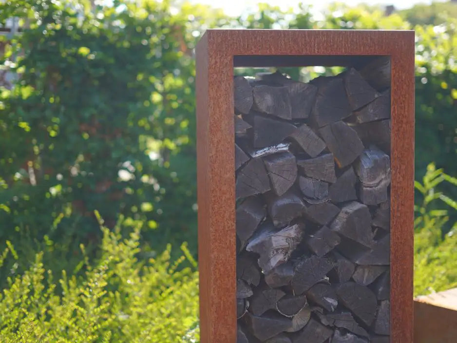 Ein schmales Cortenstahl Holzlager vor einer grünen Hecke