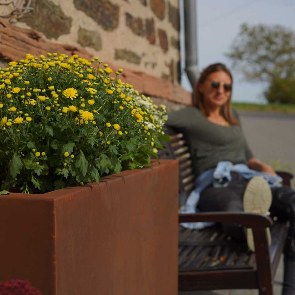 rechteckiger Cortenstahl Pflanzkübel mit gelben Pflanzen darin und einer Frau die dahinter sitzt