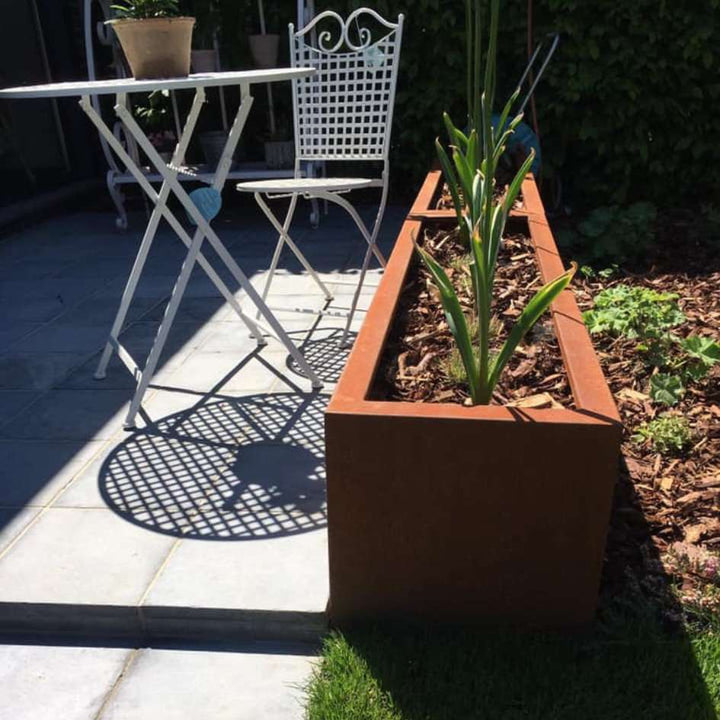 Zwei Cortenstahl Pflanzkübel hintereinander neben einer weißen Garten Sitzgarnitur
