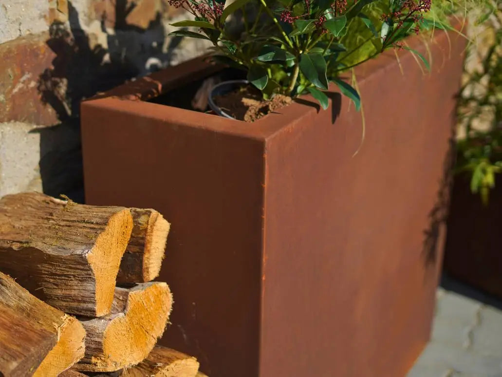 Cortenstahl Pflanzkübel mit einer grünroten Pflanze darin und einem Holzstapel davor